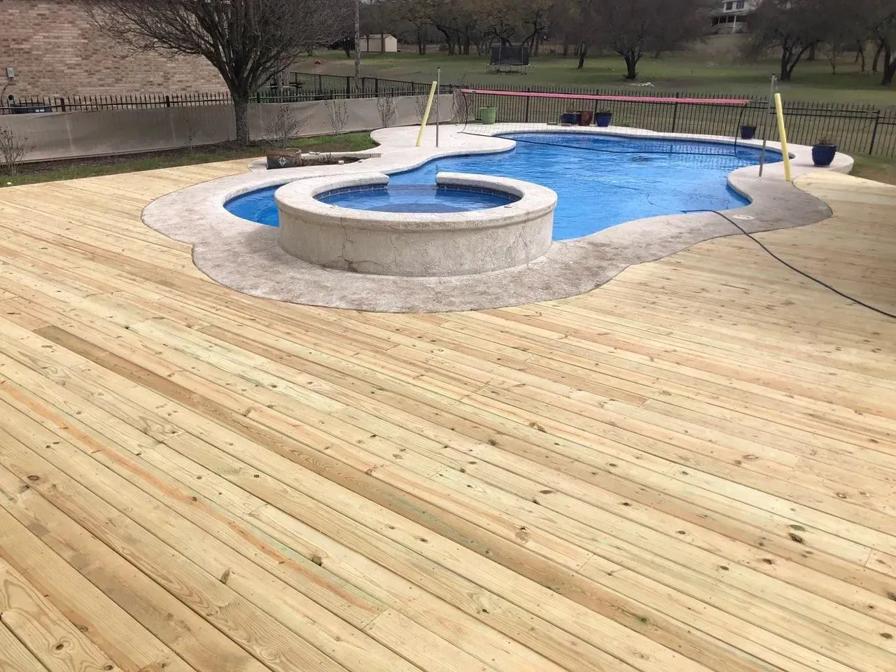 Wooden deck surrounds a freeform pool and hot tub in a backyard setting.