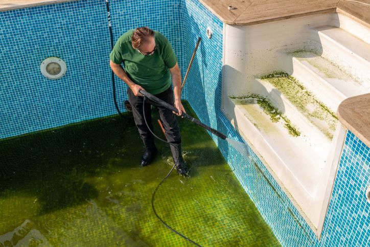 Person pressure-washing a green pool deck beside blue-tiled steps and a swimming pool