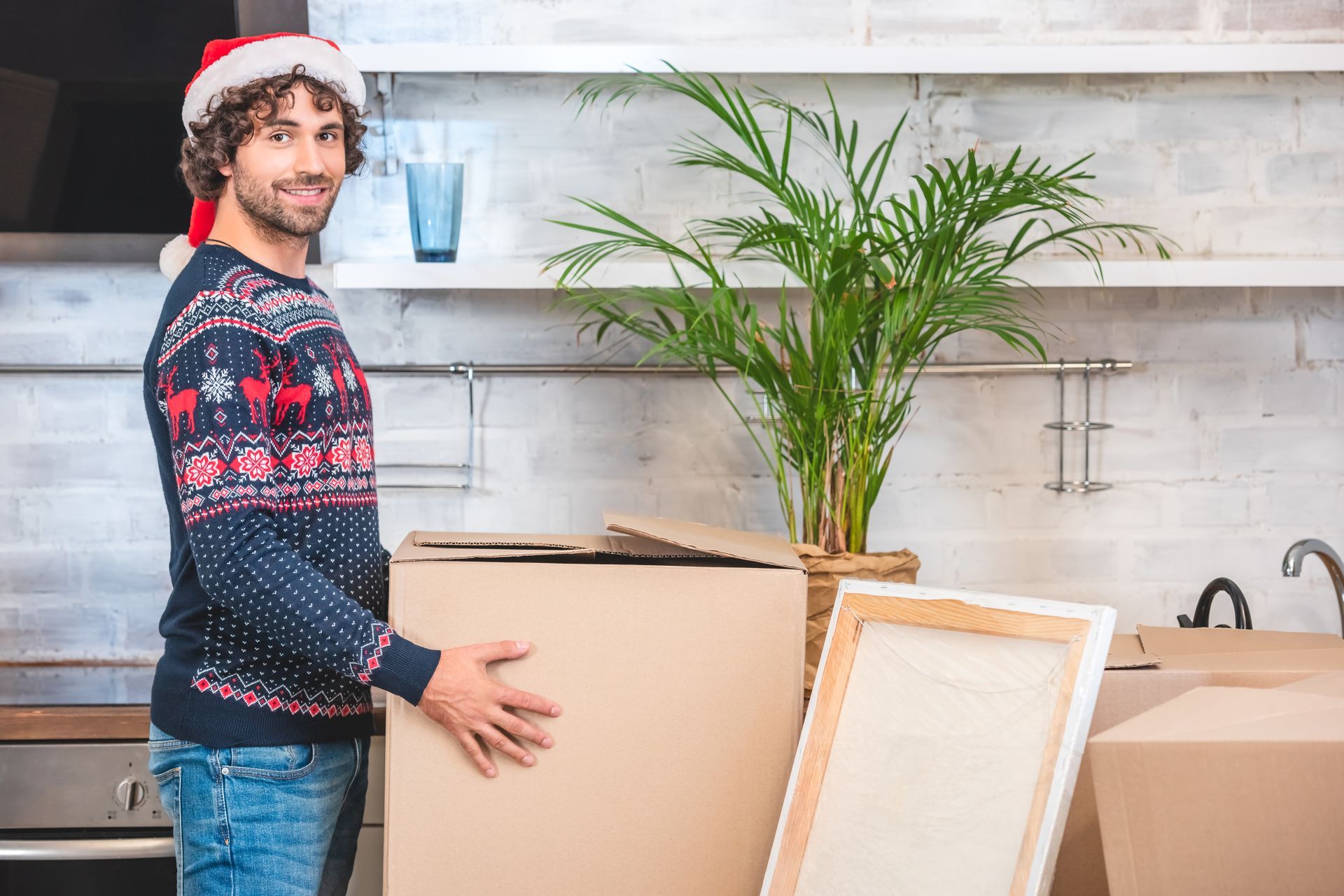Man in Christmas sweater and hat, holding a moving box in a kitchen, smiling.