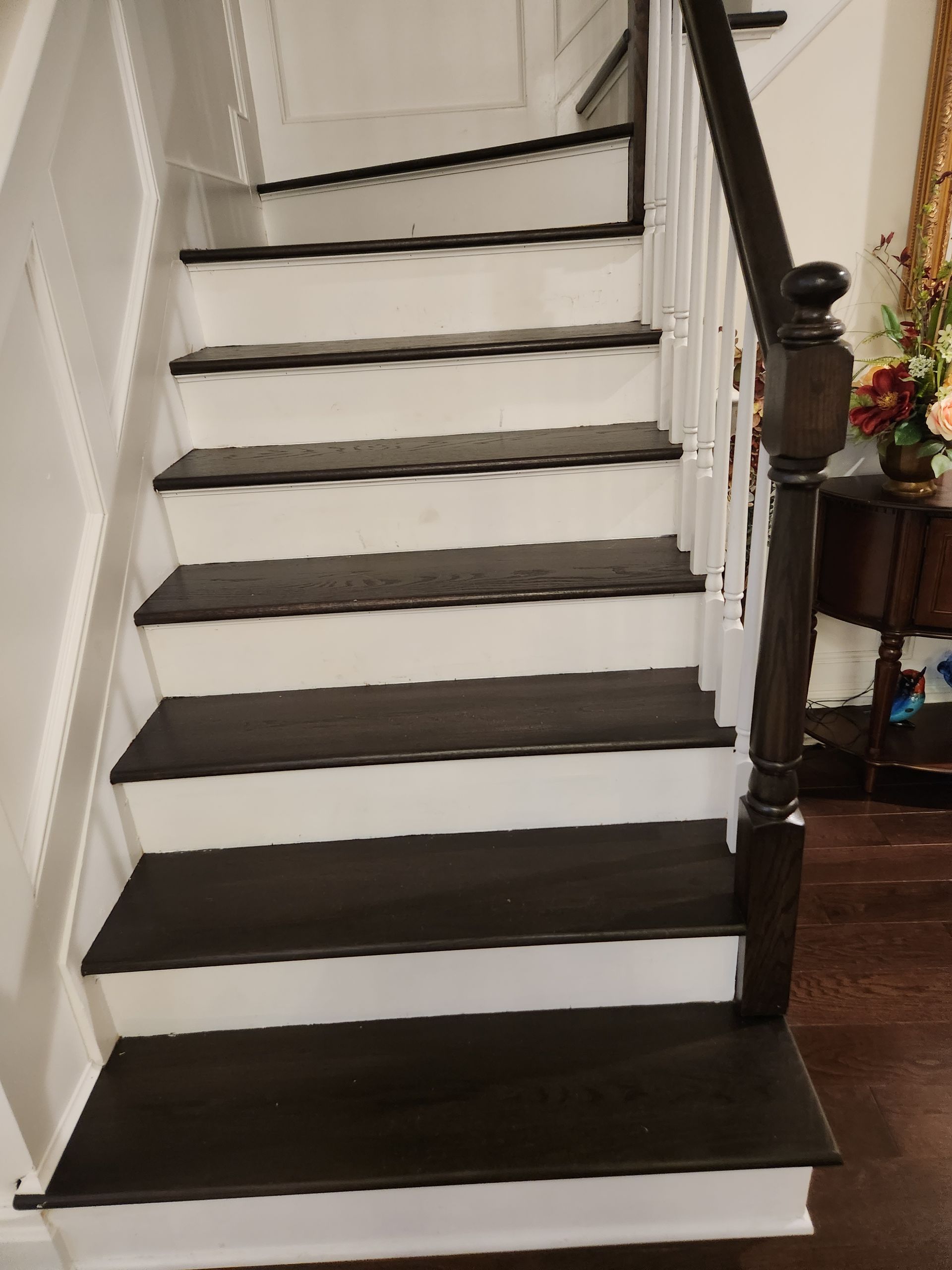 Staircase with dark treads and white risers, dark handrail. The setting is indoors, with a wall and a table visible.