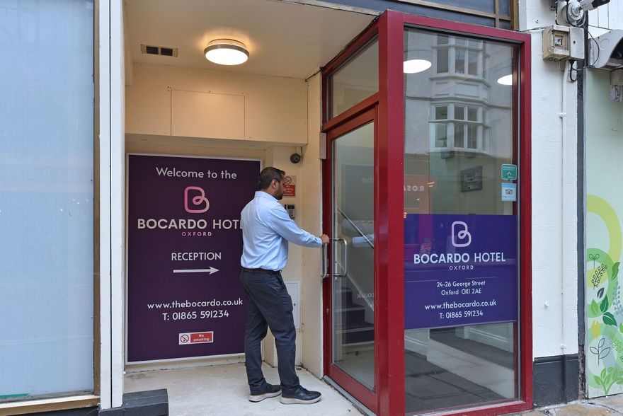 A person in a light blue shirt and dark trousers opens the red-framed glass door to the Bocardo Hotel, Oxford.