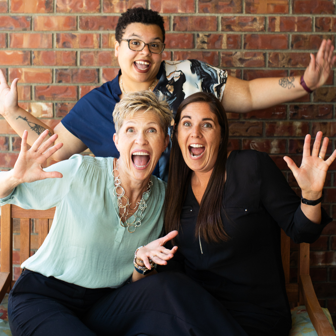 Three people with excited expressions, arms raised, in front of a brick wall.