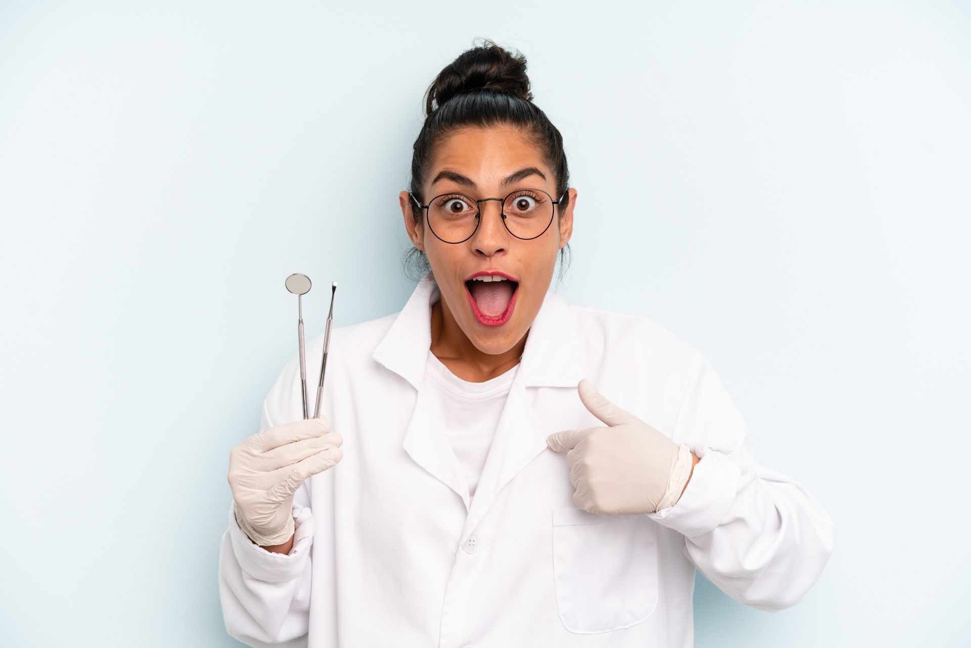Woman in lab coat, glasses, and gloves, holding dental tools, looks surprised and points at herself.
