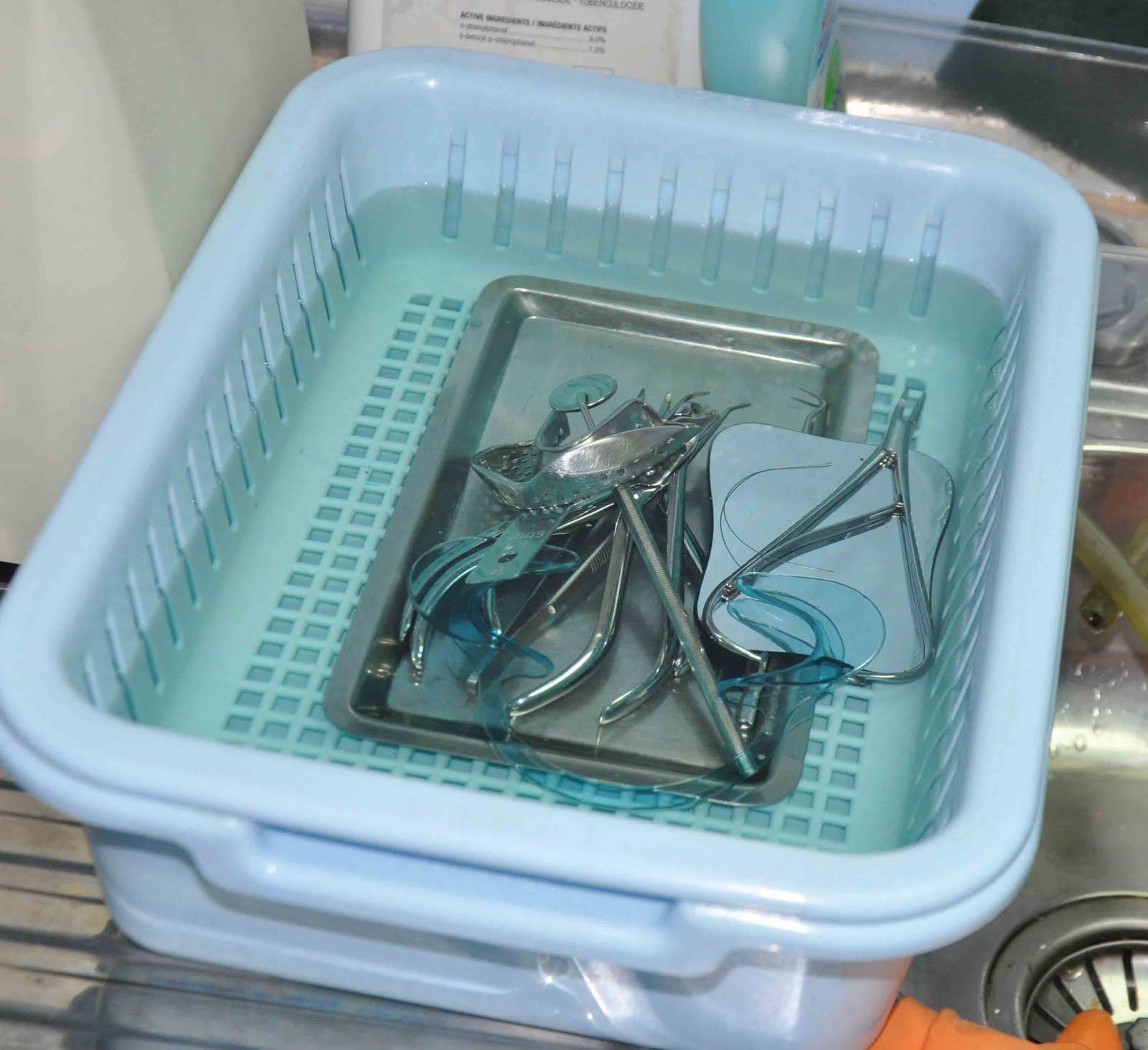 Surgical tools submerged in a blue liquid within a light blue basket, placed in a sink.
