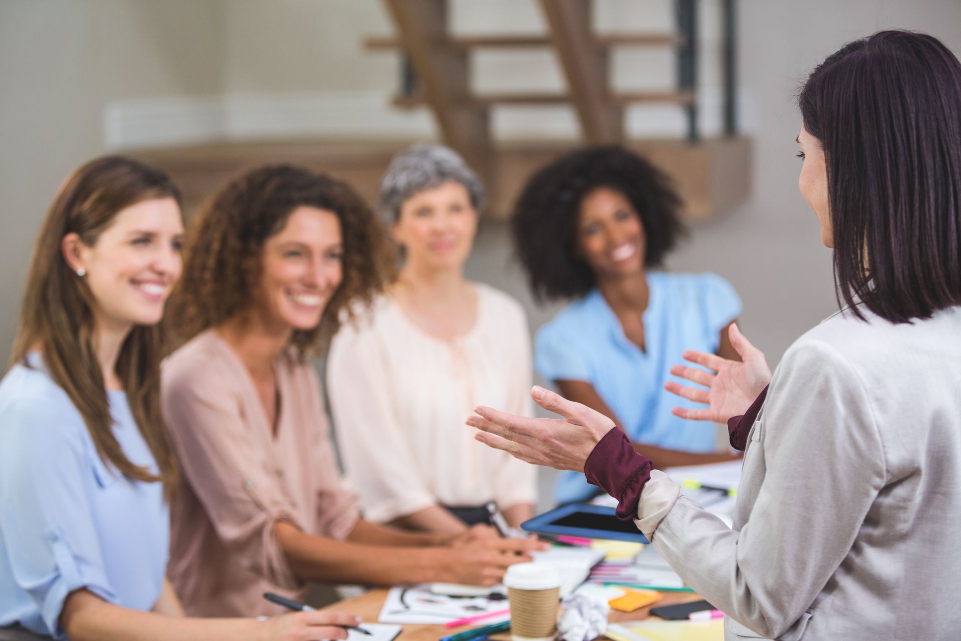 A woman gestures, speaking to a group of smiling women at a table in a bright room.