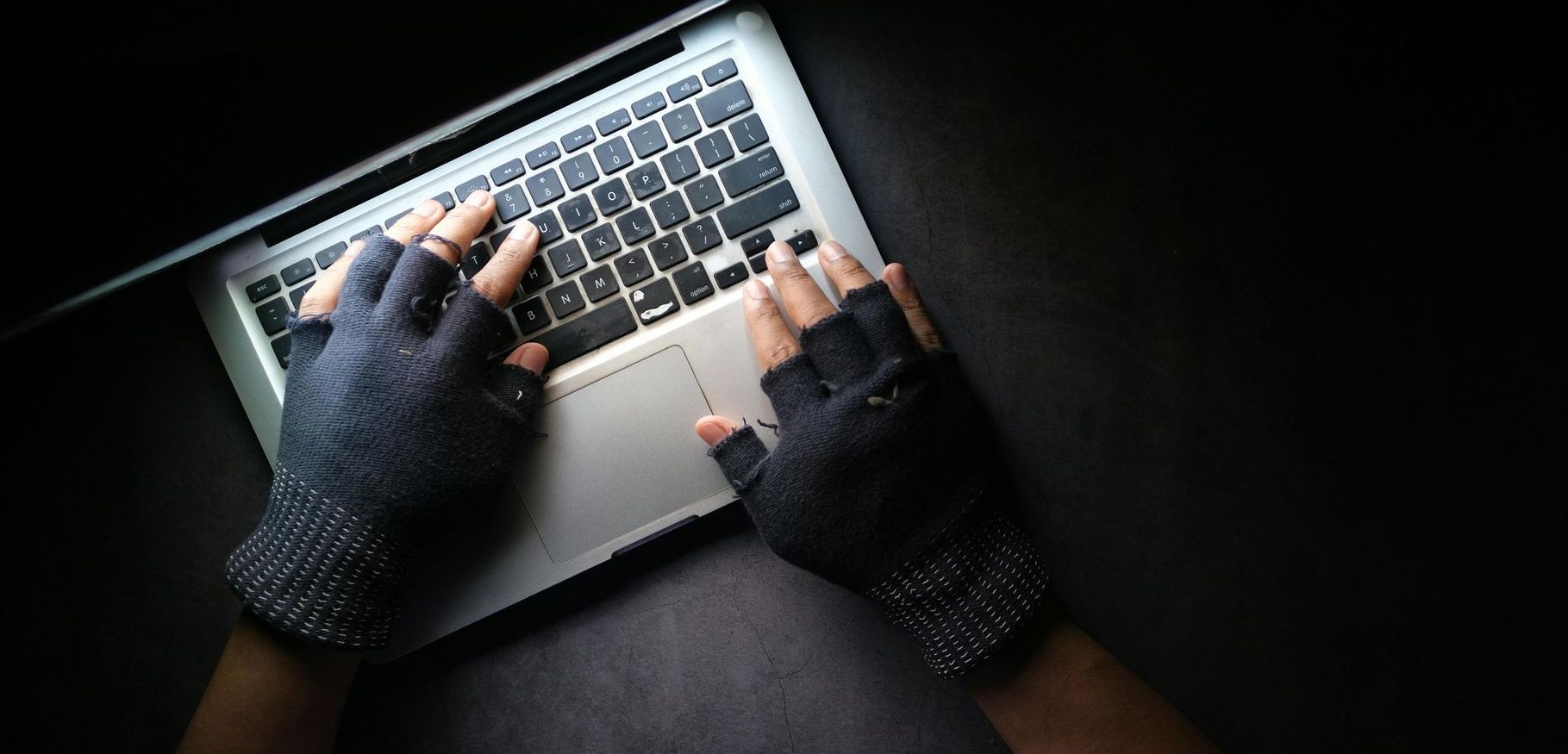 Hands wearing fingerless gloves typing on a laptop in the dark.