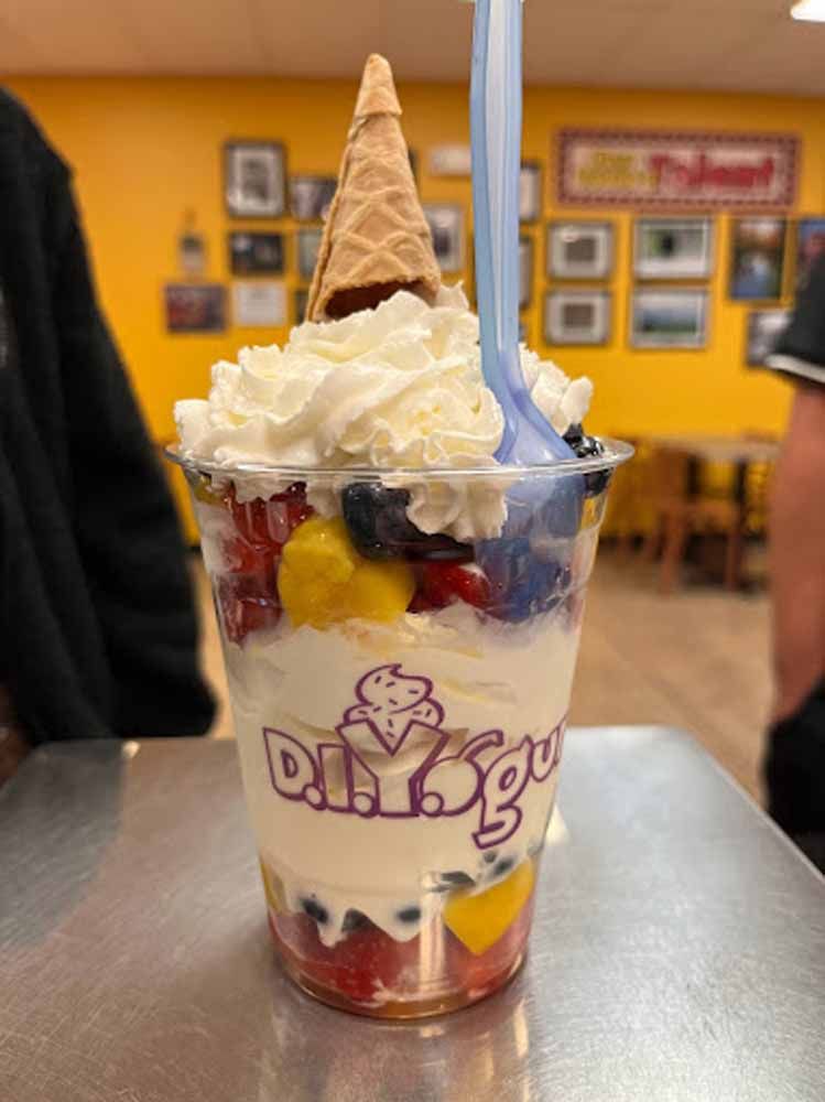 Soft-serve sundae in a clear cup with whipped cream, waffle cone, fruit, and purple logo on a table