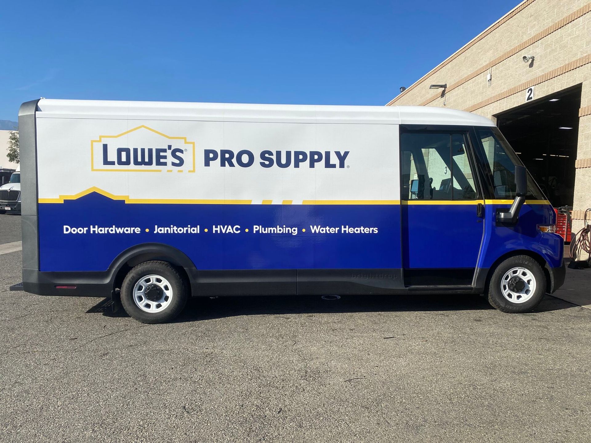 A white and blue Lowe's Pro Supply delivery van parked on a gravel lot near a brick building.