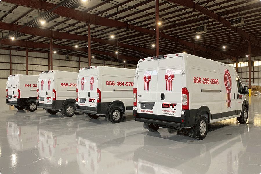 Four white cargo vans parked in a row inside a large, industrial warehouse with a shiny floor.