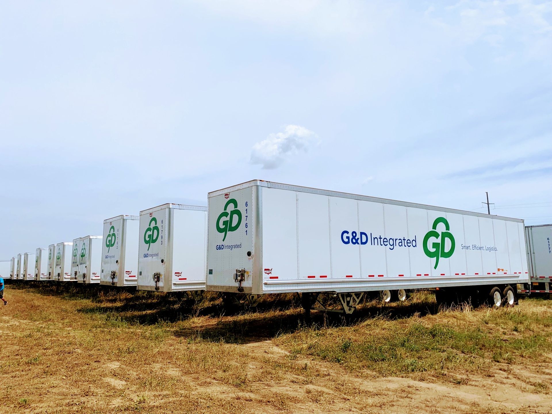 A row of white G&D Integrated semi-trailers parked in a dry, grassy field under a bright, cloudy sky.