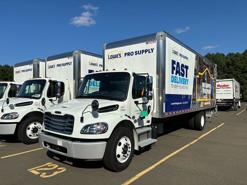 Several white Lowe's Pro Supply box trucks are parked in a paved lot under a blue sky.