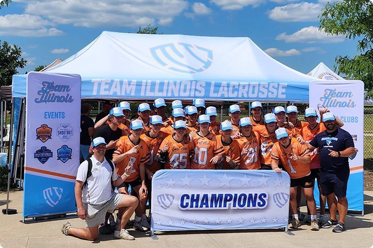 Team Illinois lacrosse players and coaches celebrating their championship victory under a branded tent outdoors.