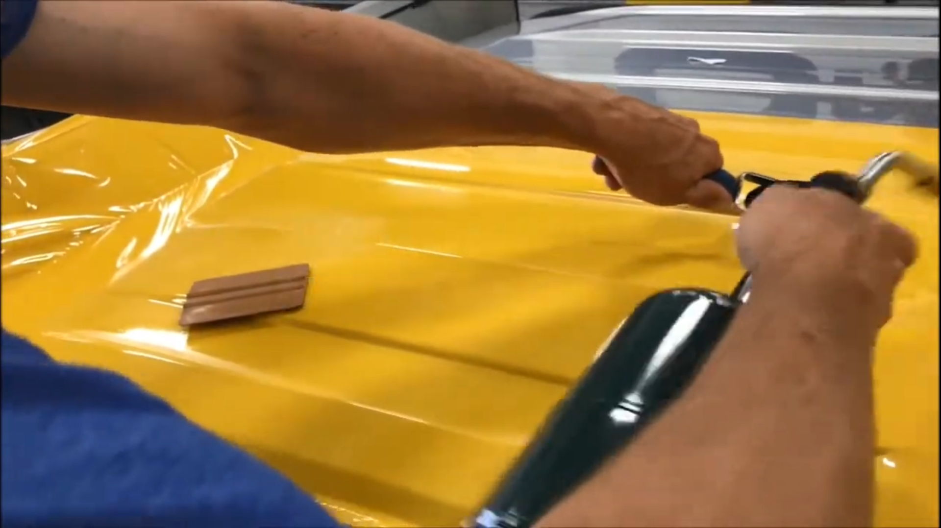 A person applies yellow automotive vinyl wrap to a vehicle surface using a heat gun and a squeegee.