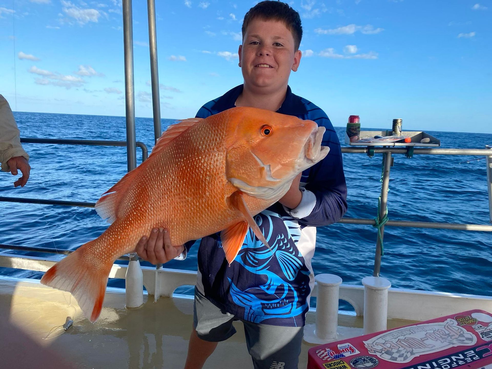A person holds a large, bright orange fish while standing on a boat deck against an ocean background under a blue sky.