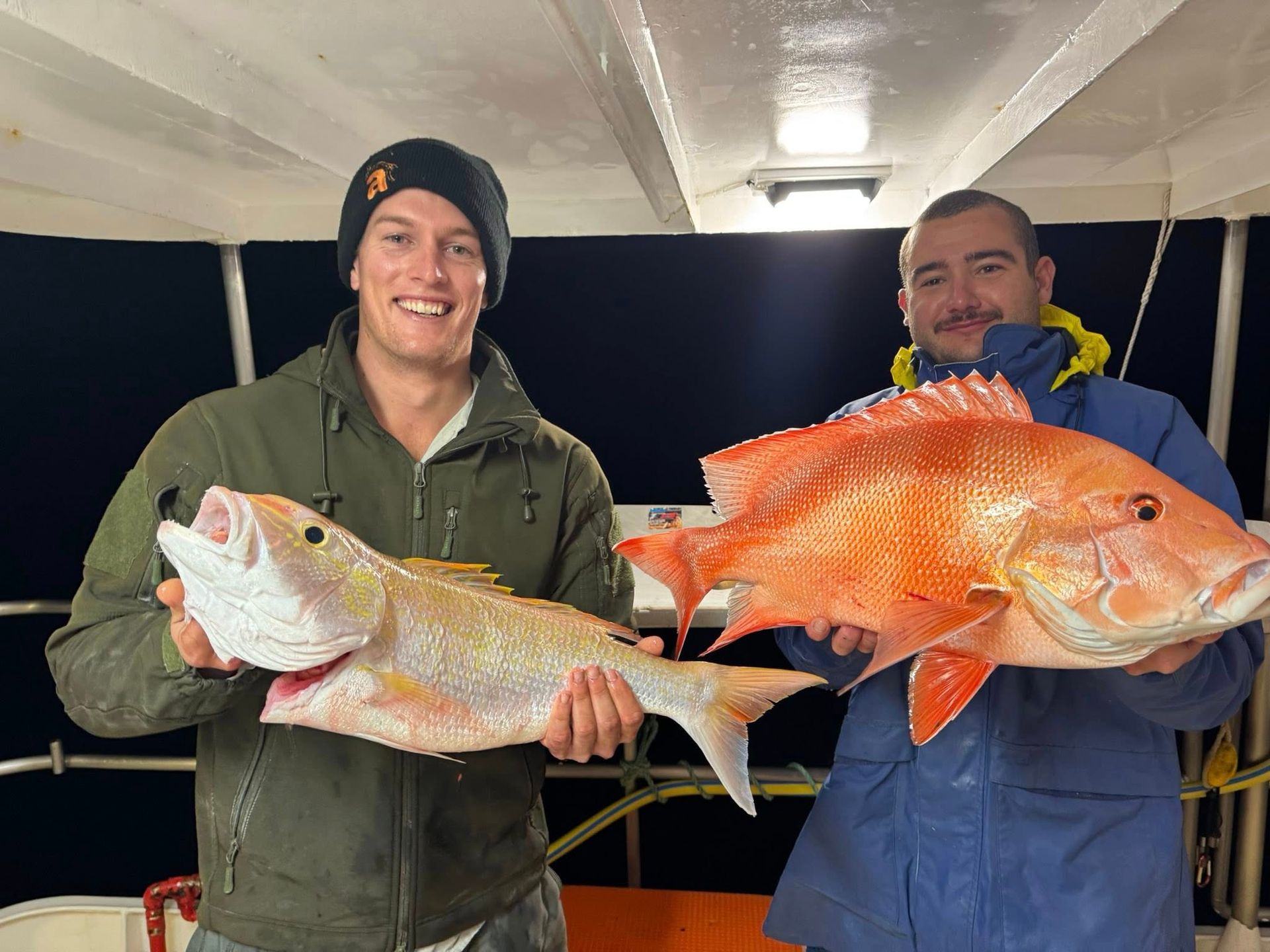 Two people smiling while holding large red fish on a boat at night.