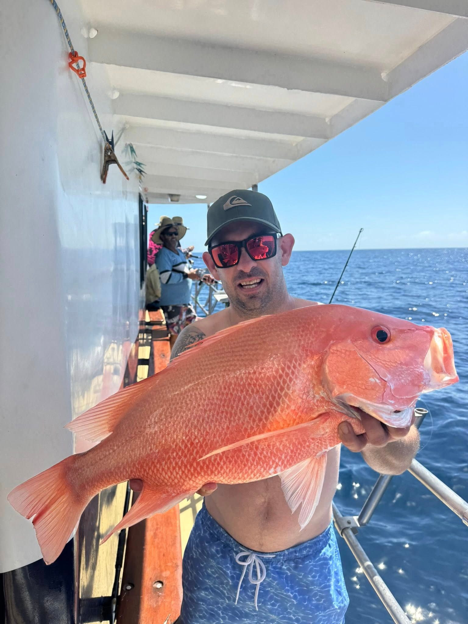 A man wearing sunglasses and a cap holds a large, bright pink fish on a boat deck with the ocean in the background.