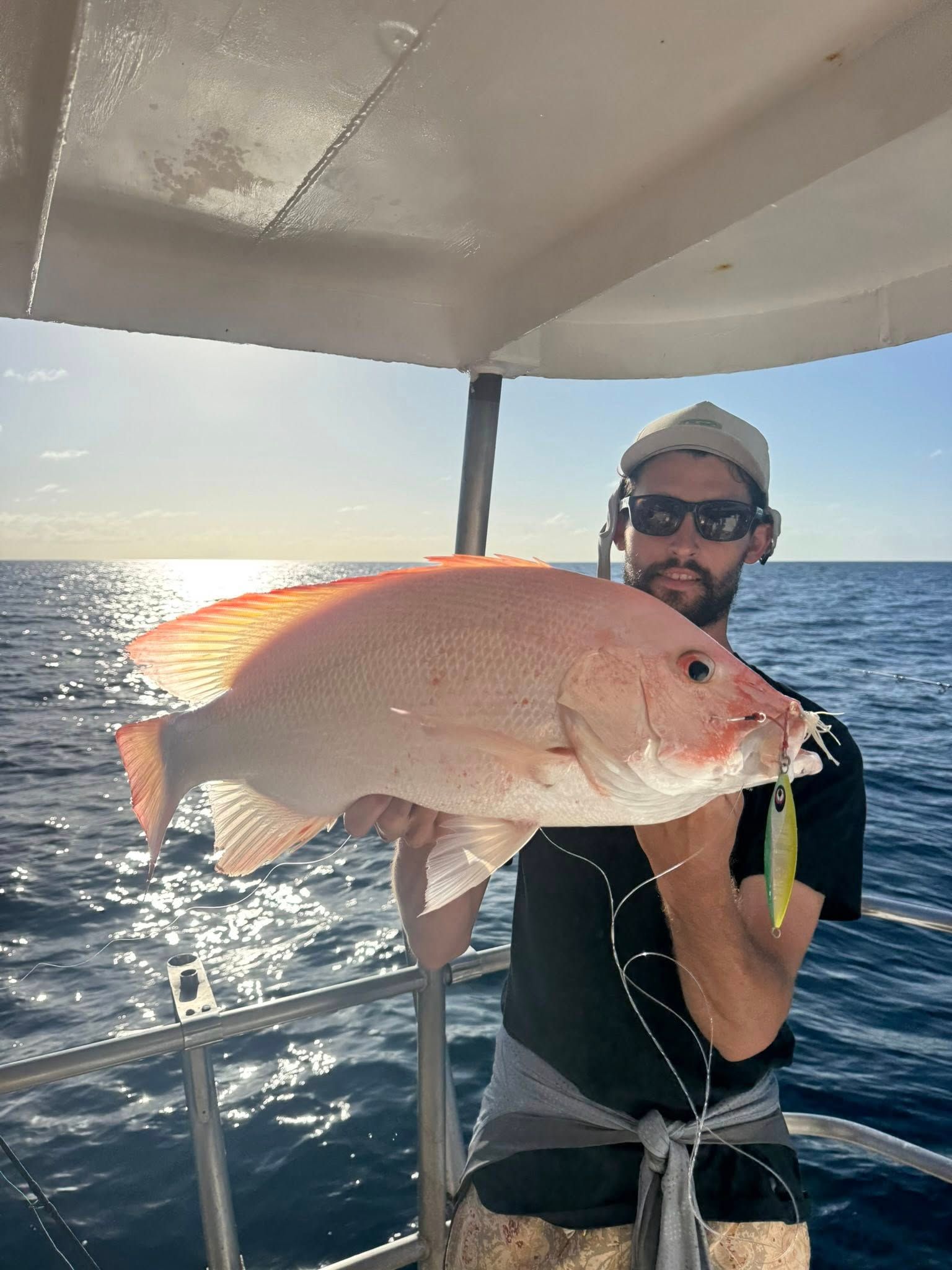 A person wearing sunglasses and a hat holding a large, pinkish fish caught on a fishing lure aboard a boat on the ocean.