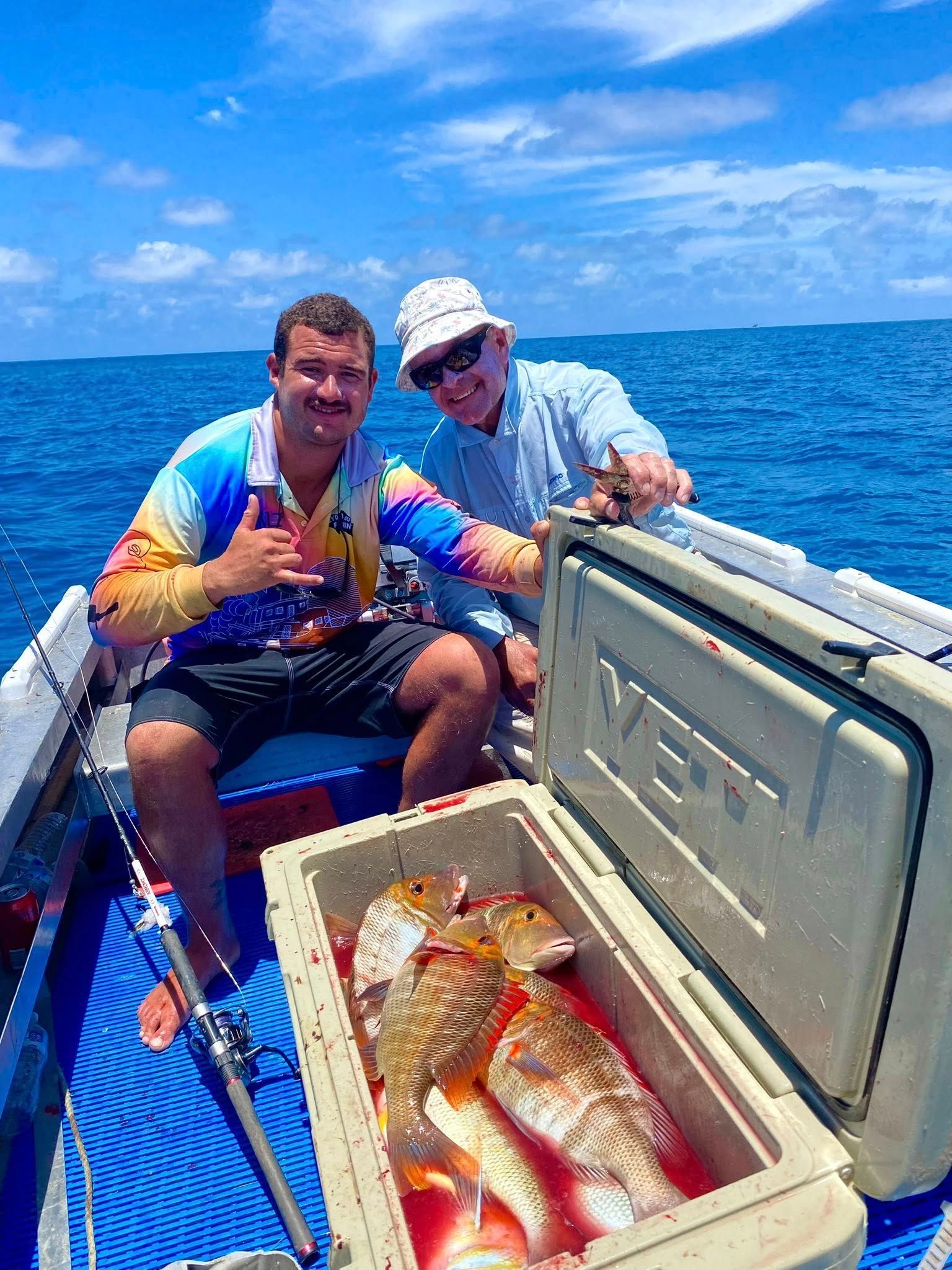Two people on a boat with a cooler full of freshly caught fish, one gesturing with a shaka sign on a sunny day.