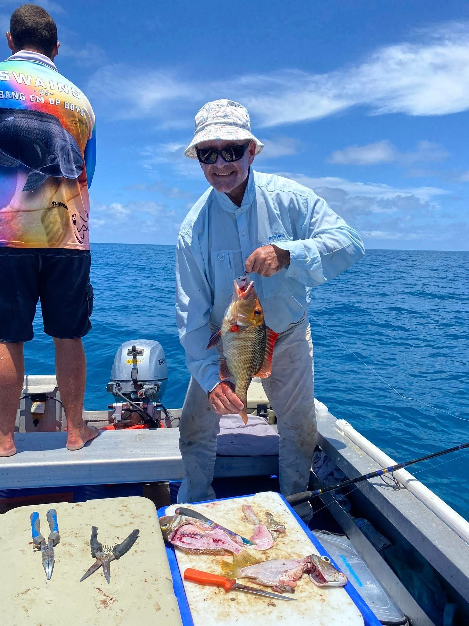 A person on a boat holds a small, colorful fish, with fishing tools on a cutting board in the foreground.