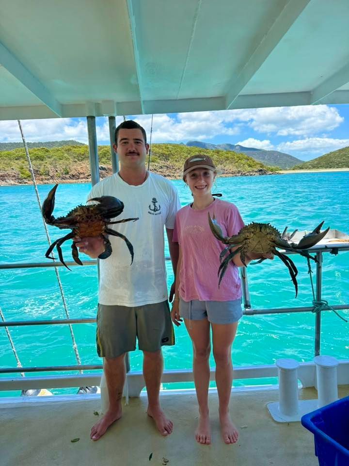 Two people stand on a boat deck holding large mud crabs, with turquoise water and a coastal landscape in the background.