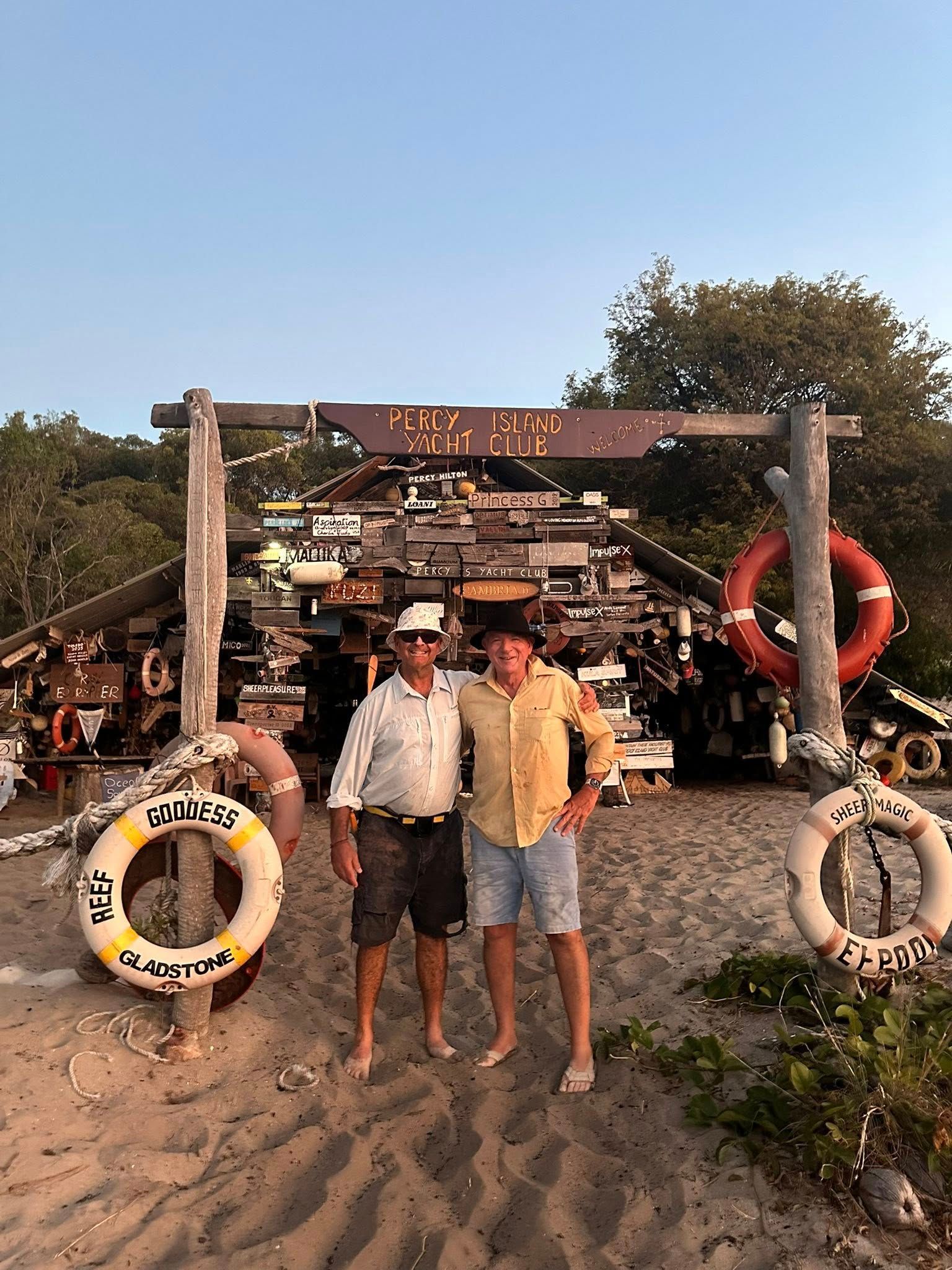 Two people stand on a sandy beach under a wooden structure decorated with lifebuoys and signage at sunset.