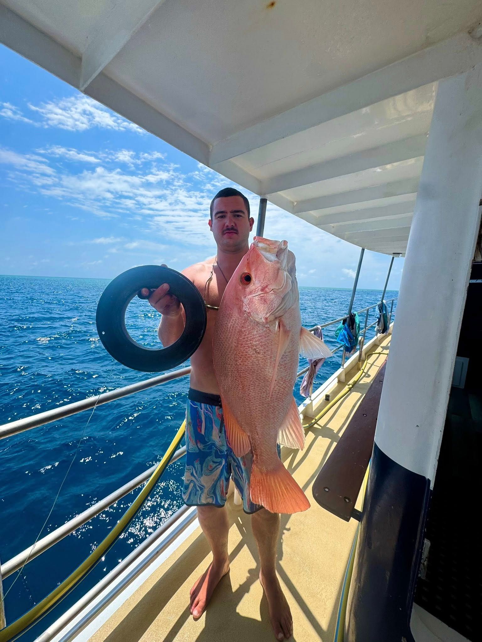 A person stands on a boat deck holding a large red snapper fish and a black circular object against an ocean background.