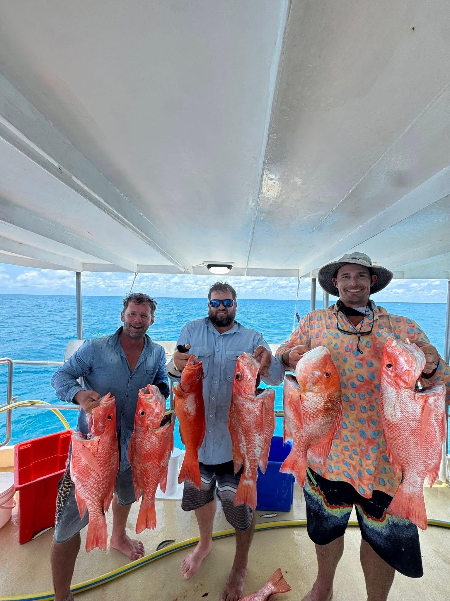 Three people on a boat deck hold up large, freshly caught red snapper fish against a bright, sunny ocean background.