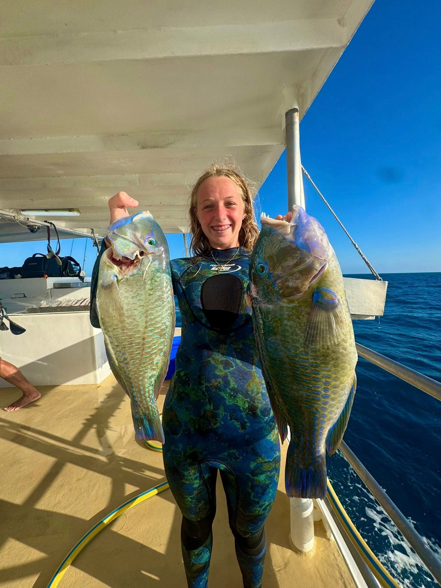 A person in a patterned wetsuit stands on a boat, smiling while holding two large, green fish on a sunny day at sea.