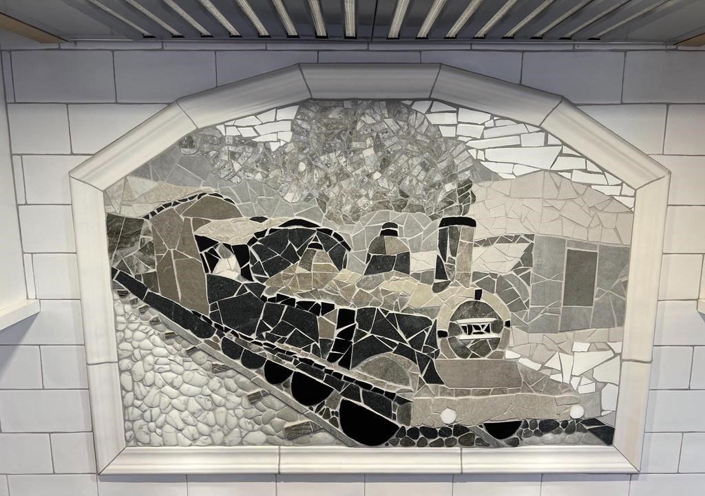 Mosaic mural of a mountainous landscape with a road and buildings, framed in a white display arch.