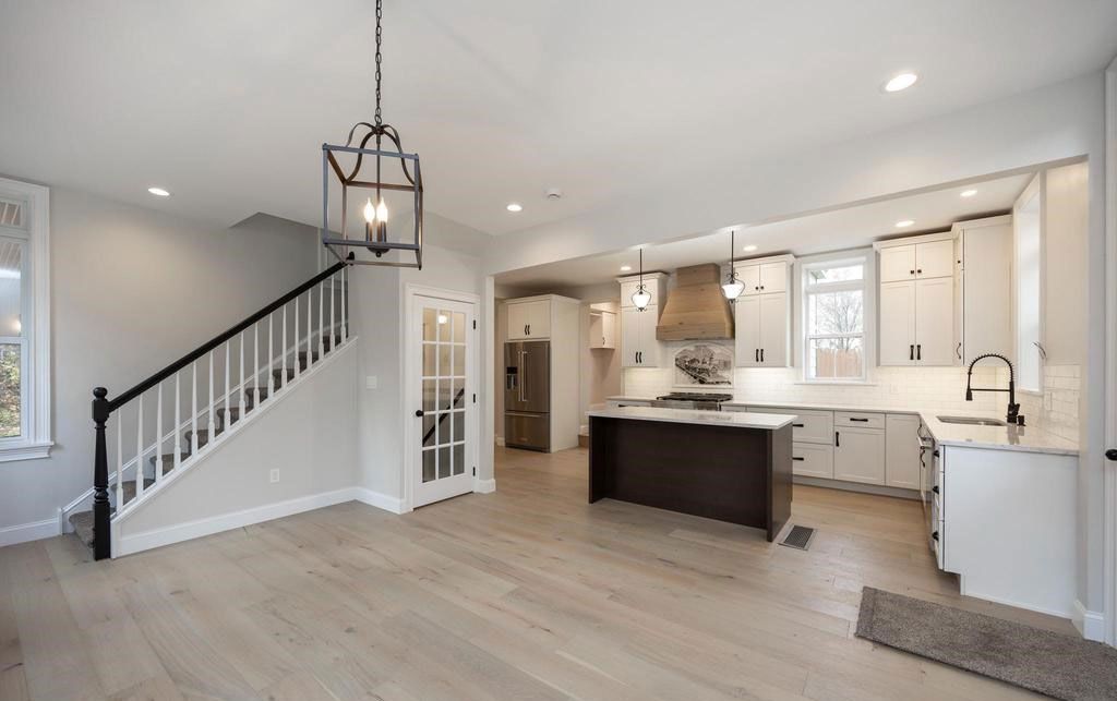 Bright open-concept kitchen and living area with staircase, island, white cabinetry, and light wood floors