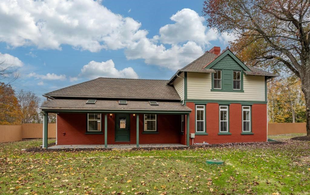 Red two-story farmhouse with a porch, green trim, and a large yard under a cloudy sky.