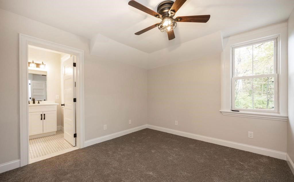 Empty carpeted bedroom with ceiling fan, closet, bathroom doorway, and window with white trim