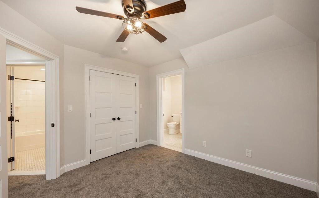 Empty beige bedroom with carpet, ceiling fan, closet doors, and an open bathroom doorway