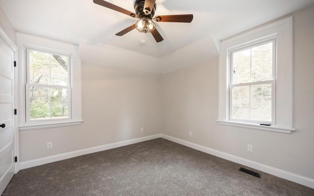 Empty beige bedroom with gray carpet, white trim, two windows, and a ceiling fan.