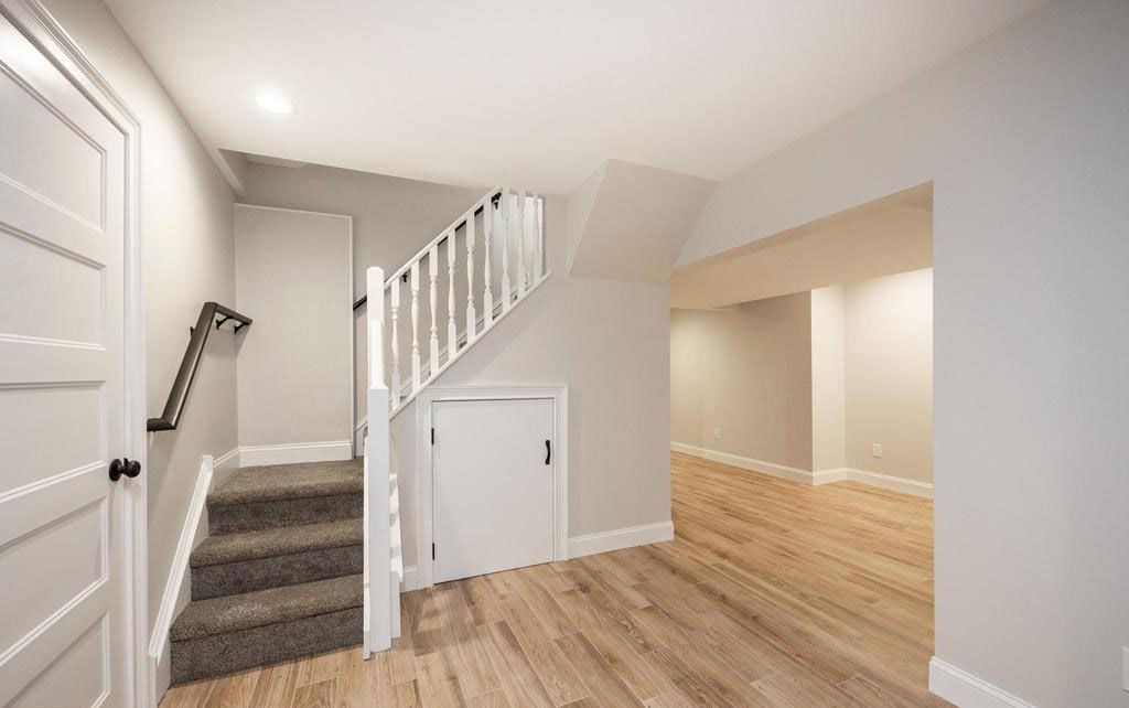 Empty hallway with hardwood floors, carpeted stairs, white walls, and a small under-stair door.