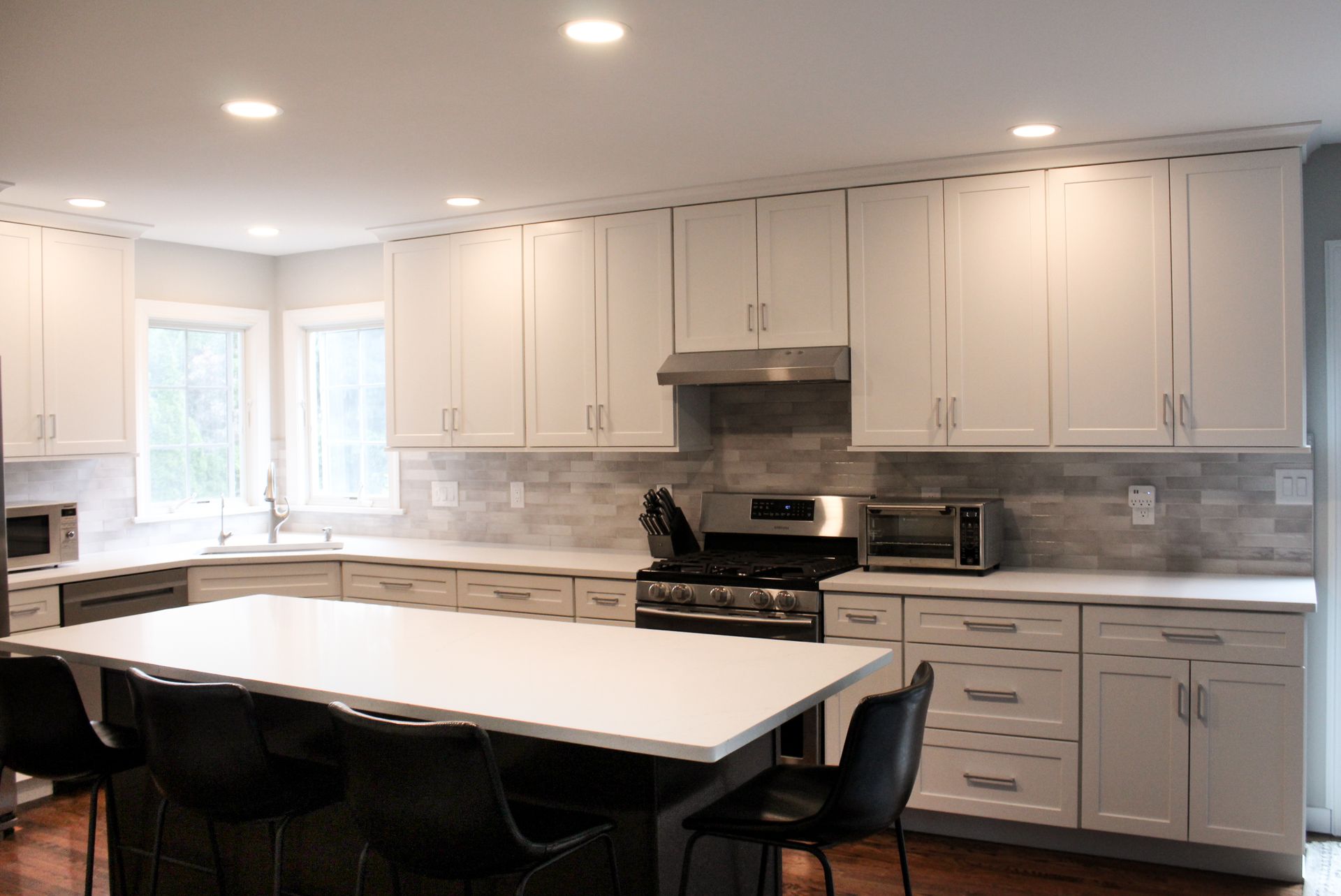 Bright modern kitchen with white cabinets, stone backsplash, island, and black chairs under recessed lights