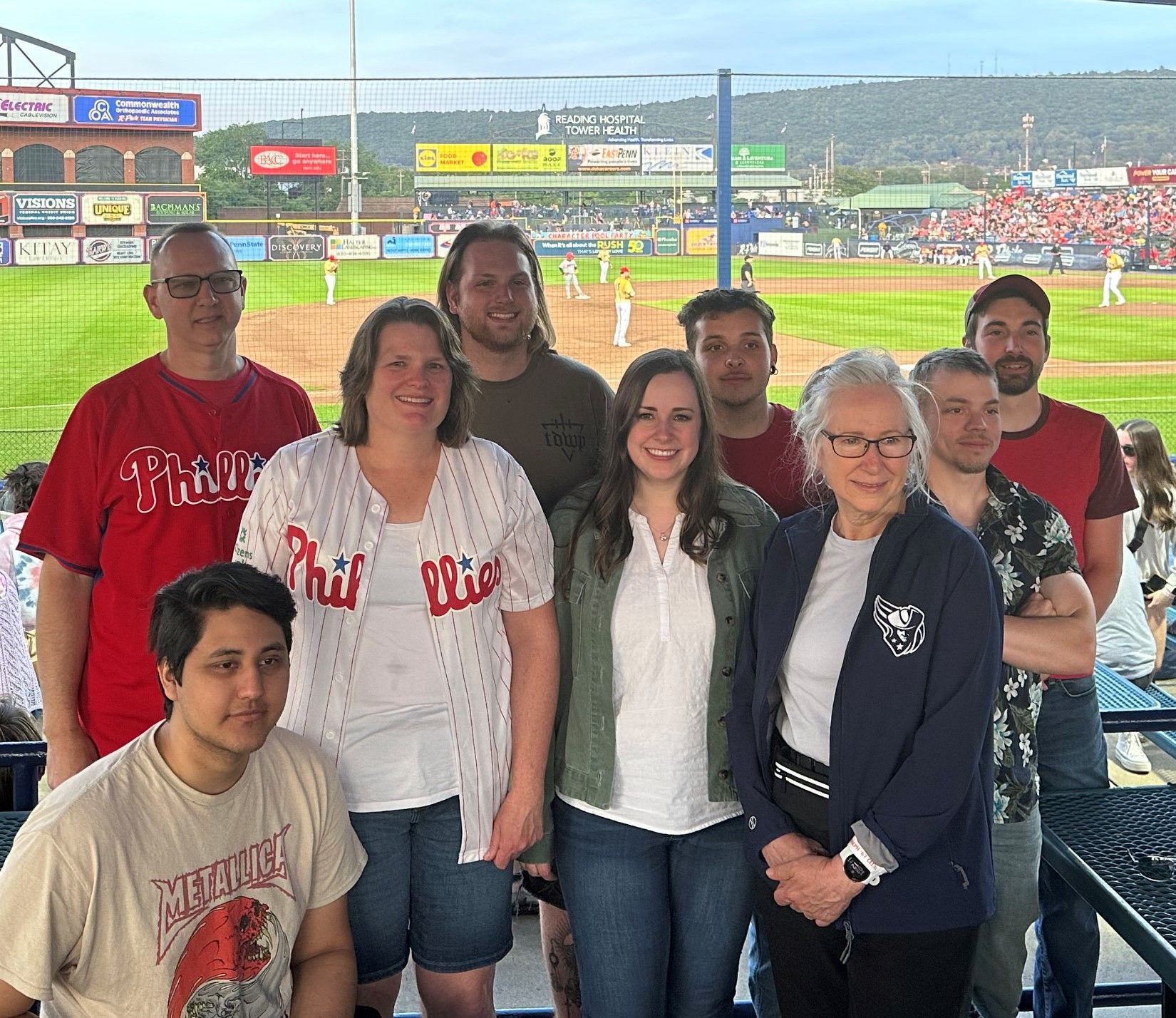 Group of fans posing at a baseball stadium, smiling in front of the field during a game.