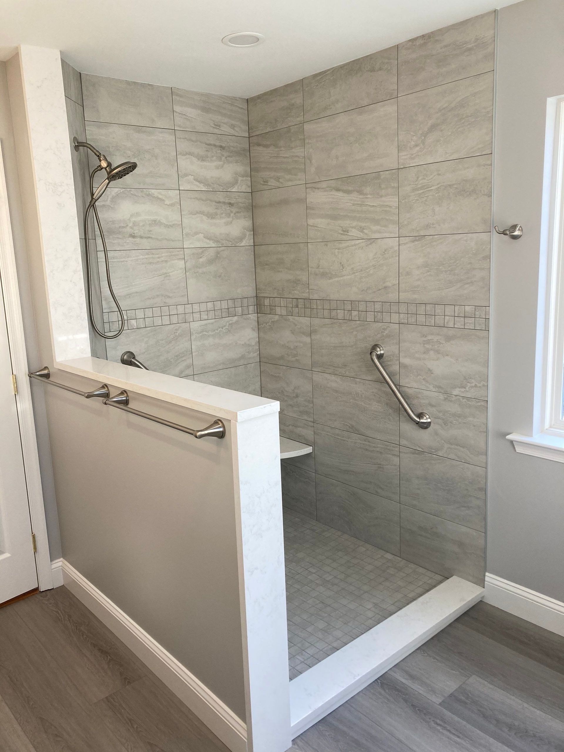 Accessible shower stall with gray tile walls, white bench, and grab bars in a bathroom