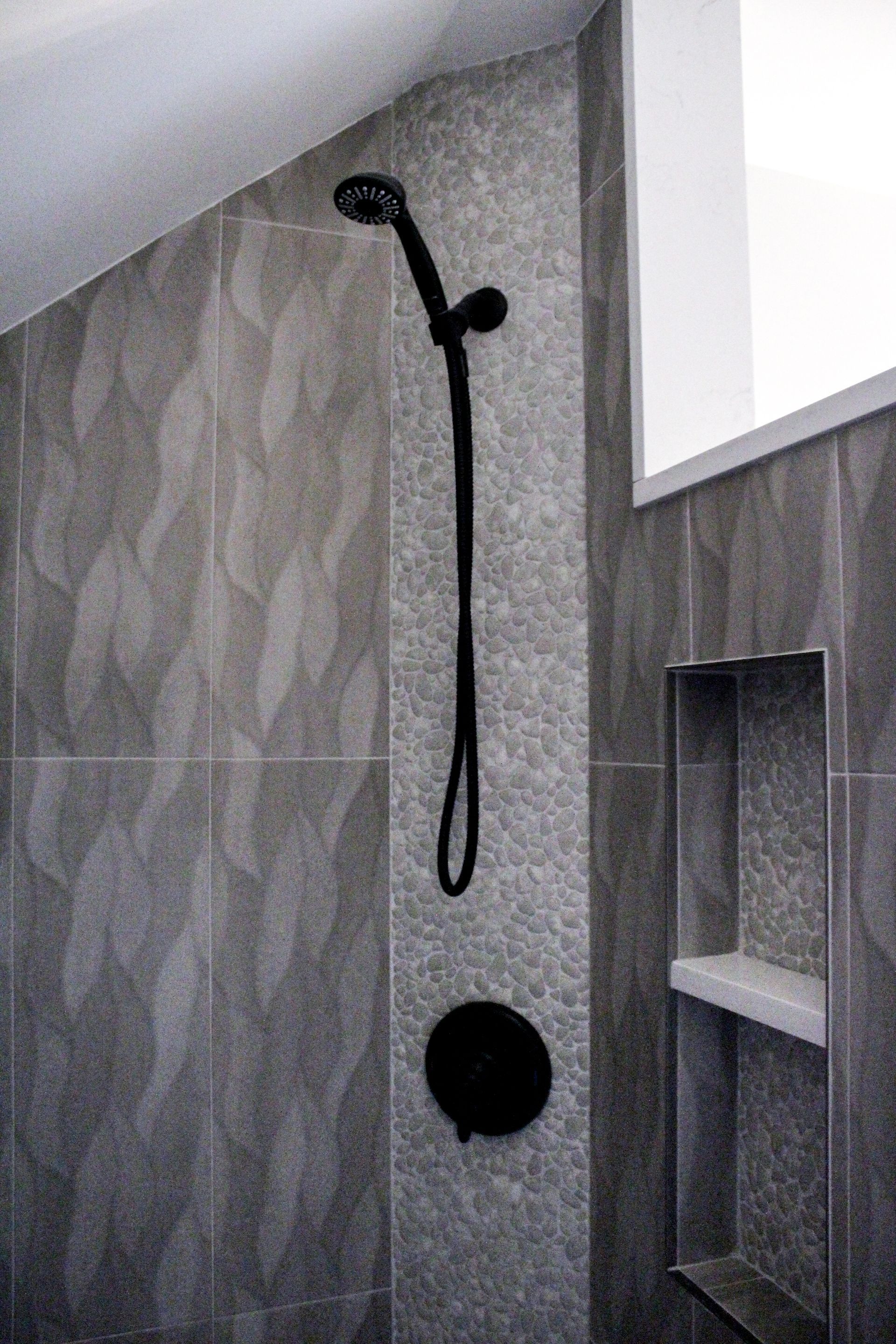 Gray tiled shower with black showerhead, hose, and recessed shelf.