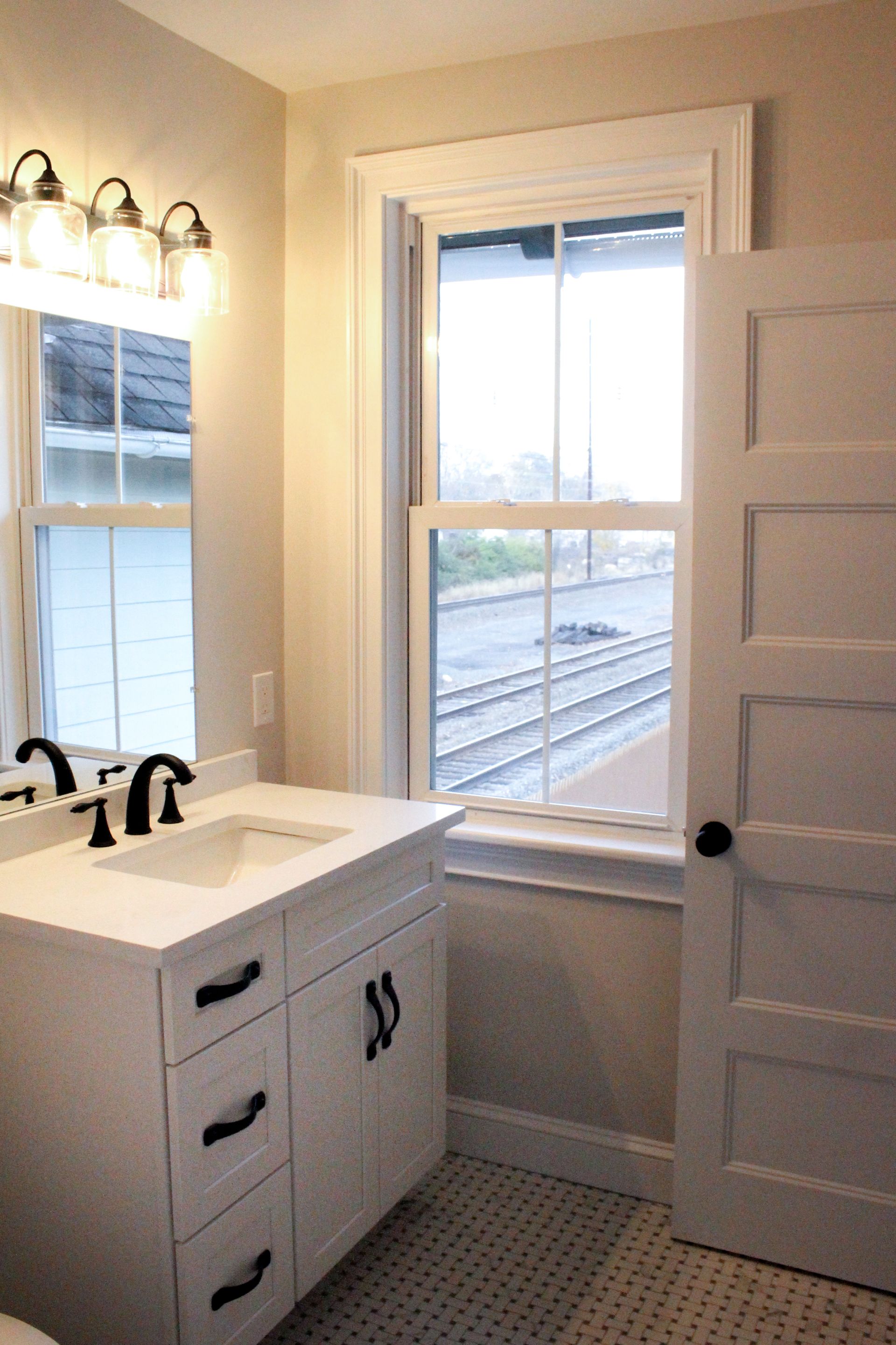 Bathroom with white vanity, sink, wall mirror lights, and a window looking onto train tracks