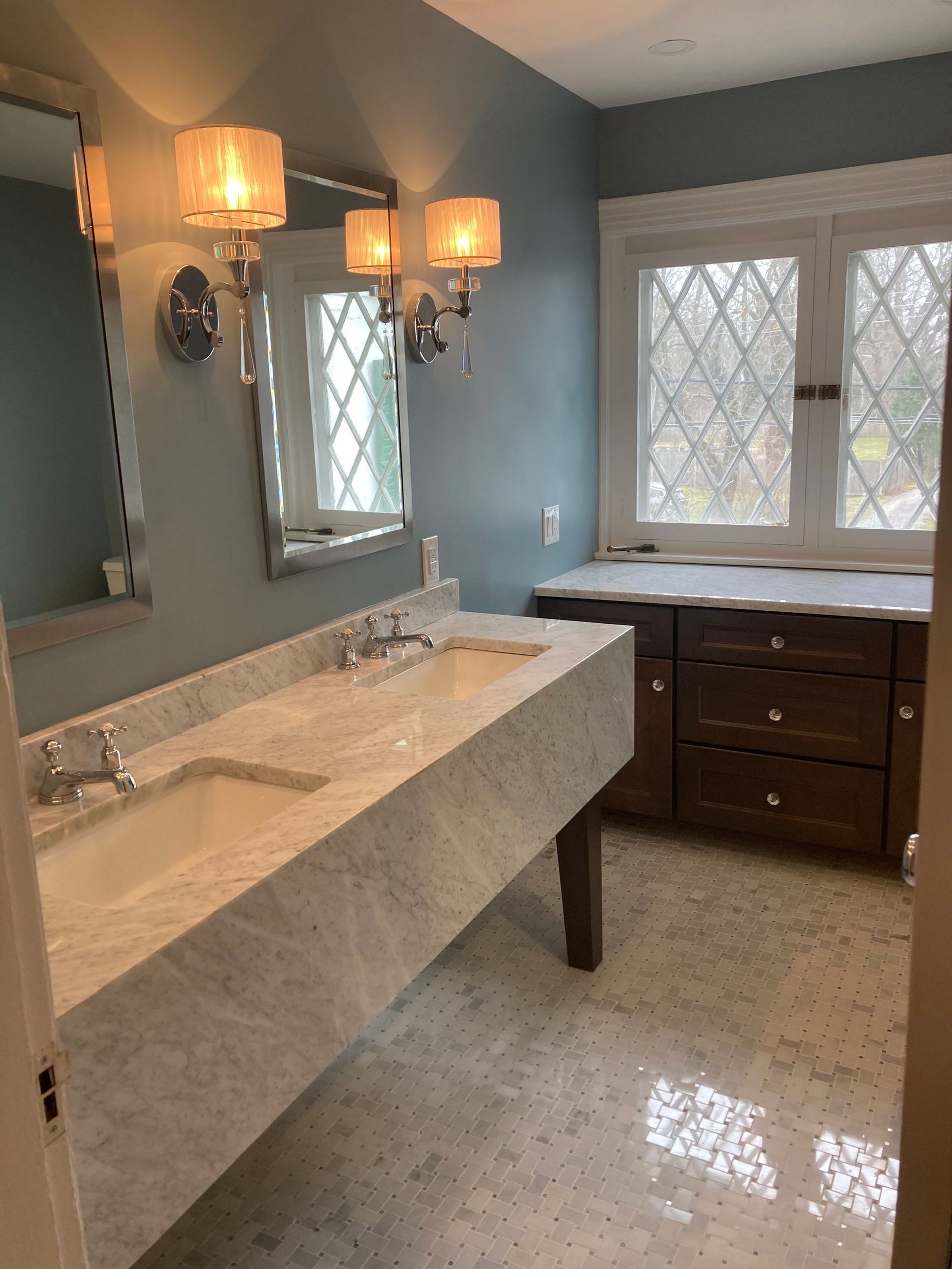 Spacious bathroom with double vanity, large mirror, and light blue walls beside a window and glass shower.