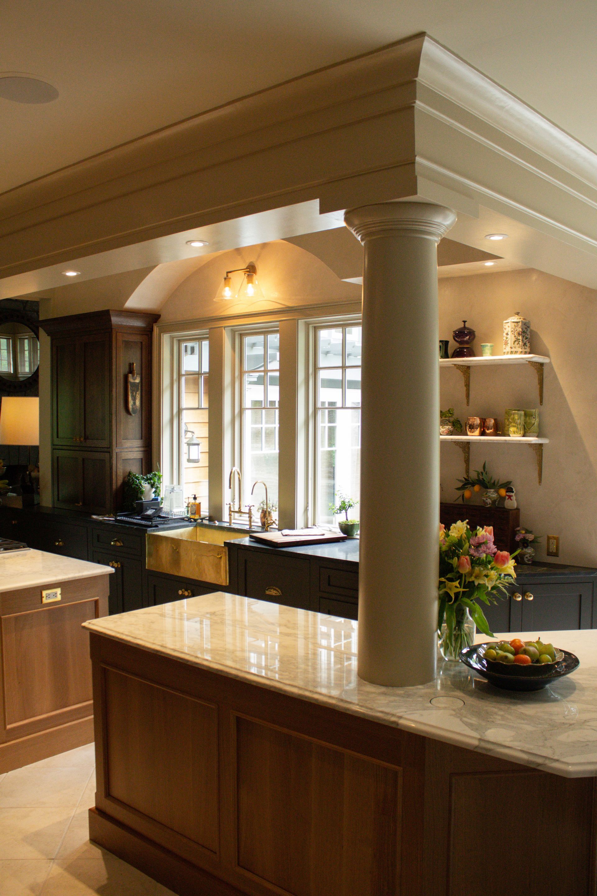 Elegant kitchen with wood cabinetry, marble island, column, and glass-front cabinets under warm lighting