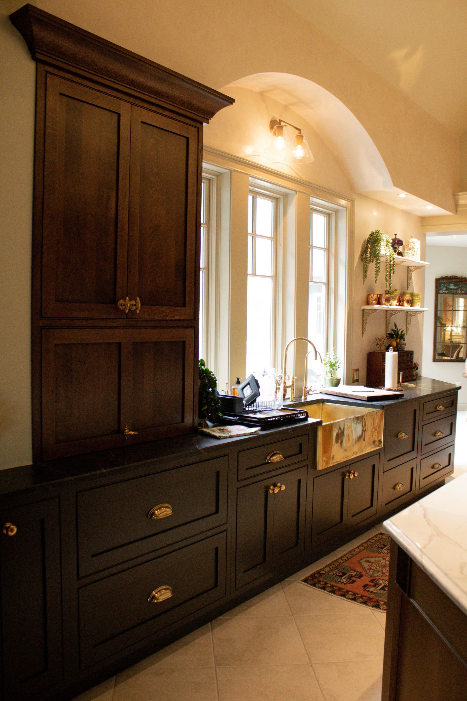 Dark kitchen with tall wooden cabinets, a sink under windows, and a patterned rug on the tile floor.