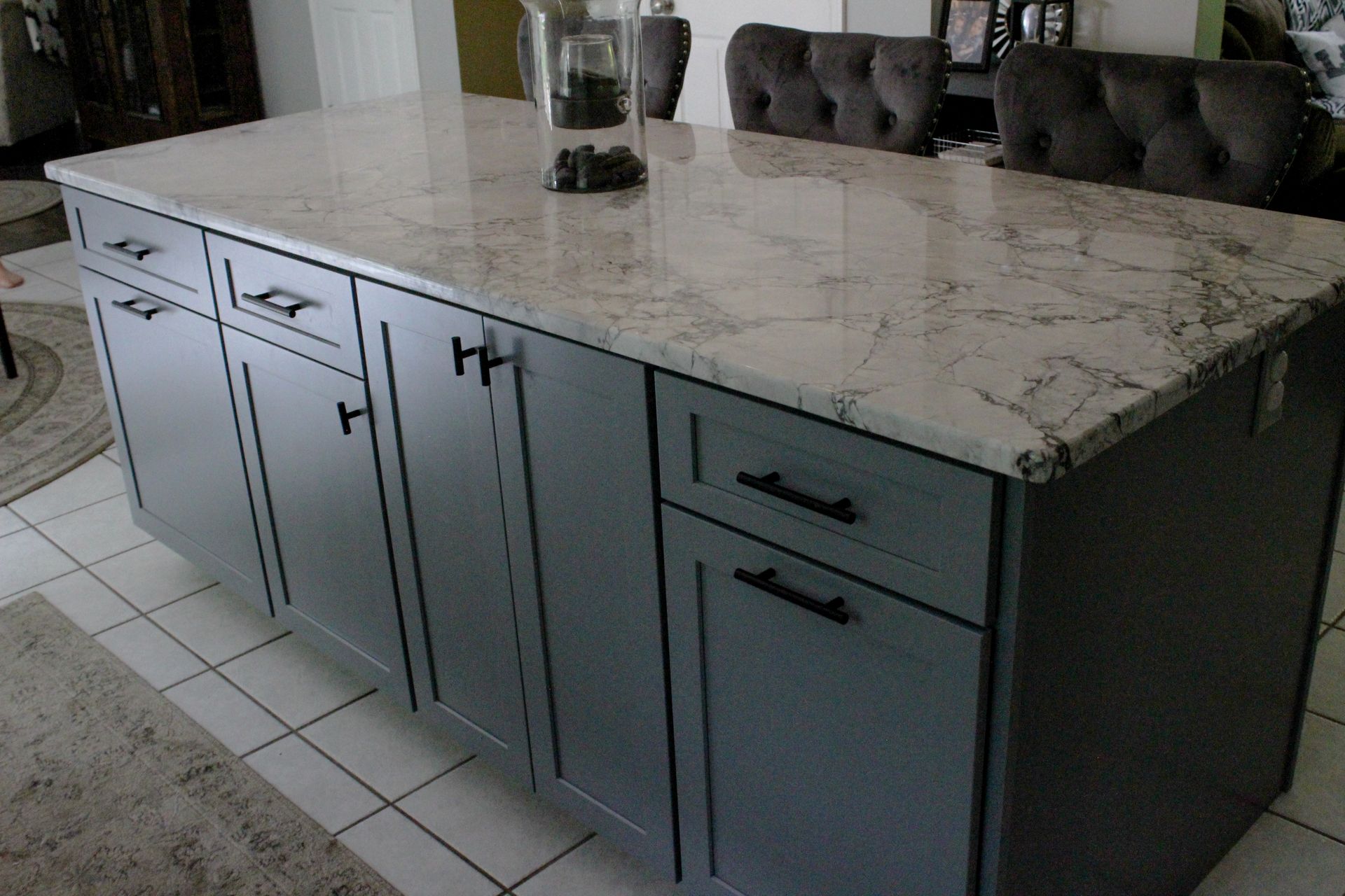 Kitchen island with white marble countertop and dark cabinet doors and drawers, viewed in a tiled kitchen