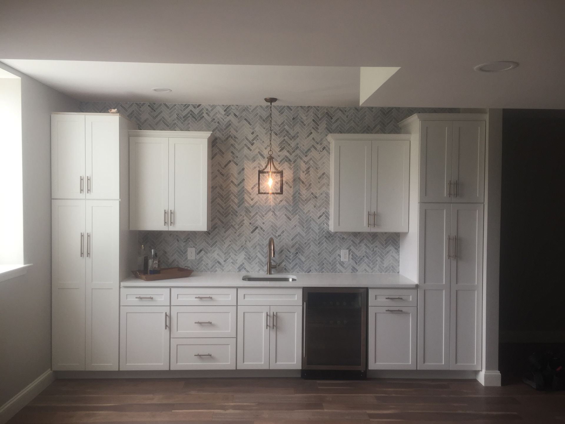 White kitchen with gray patterned backsplash, cabinets, black dishwasher, and pendant light over the sink.