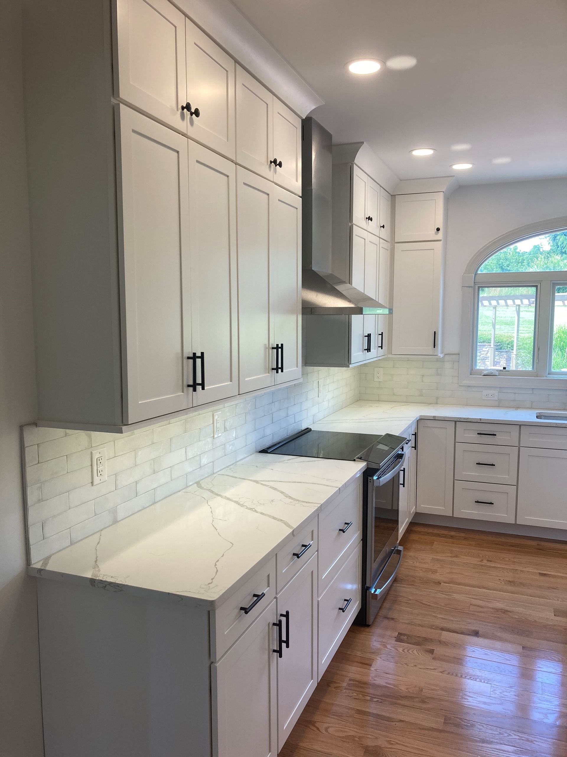 Bright white kitchen with tall cabinets, marble countertops, stainless range hood, and wood floor.