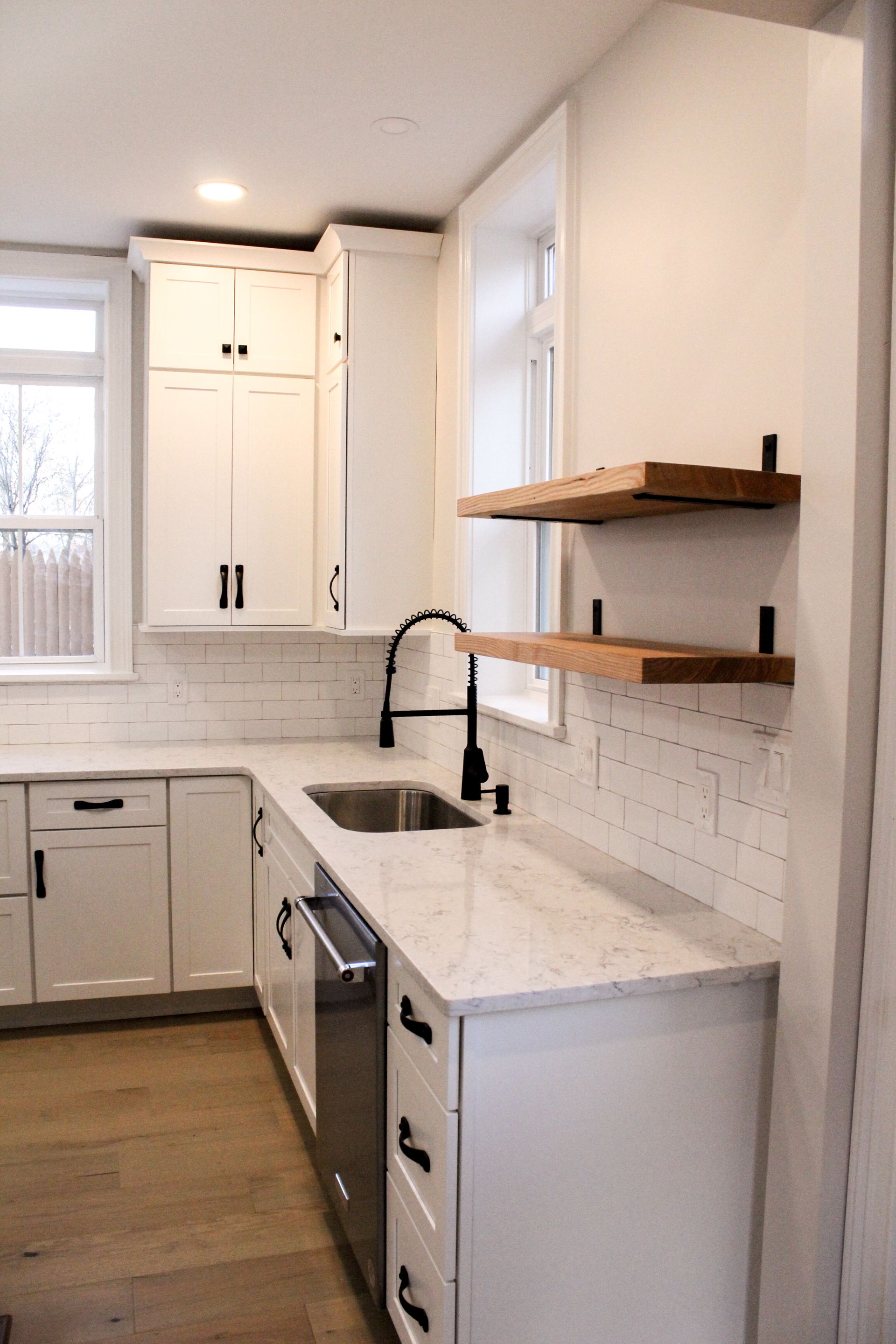 Bright white kitchen with wood shelves, black faucet, and wood floor through a narrow galley layout