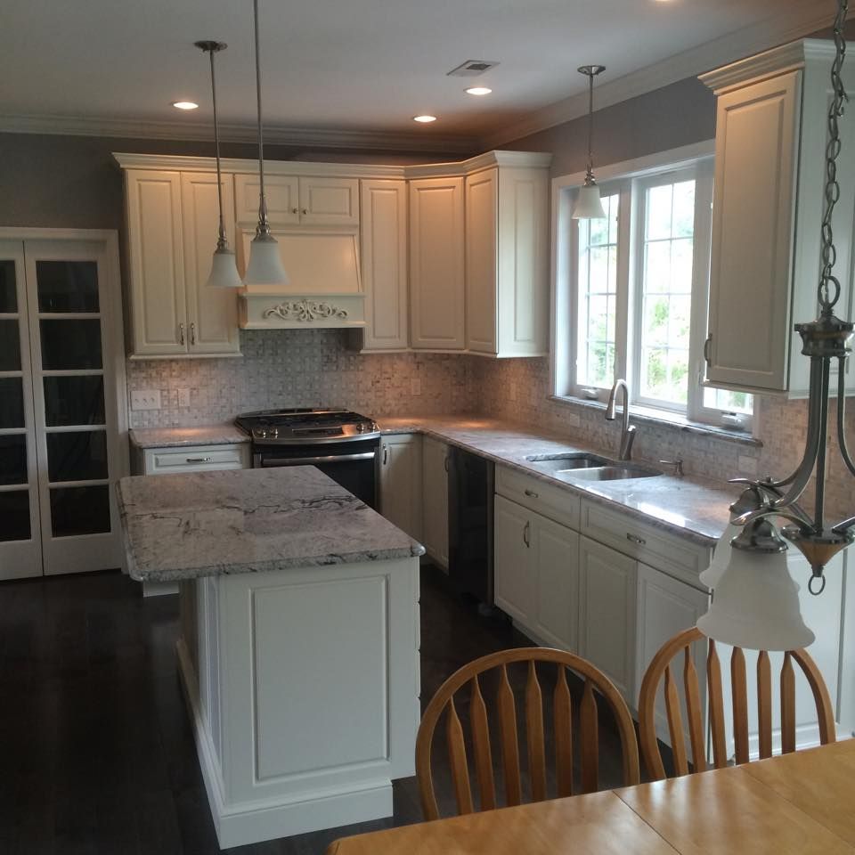 Bright kitchen with white cabinets, granite island, stainless stove, sink by window, and wood chairs in foreground