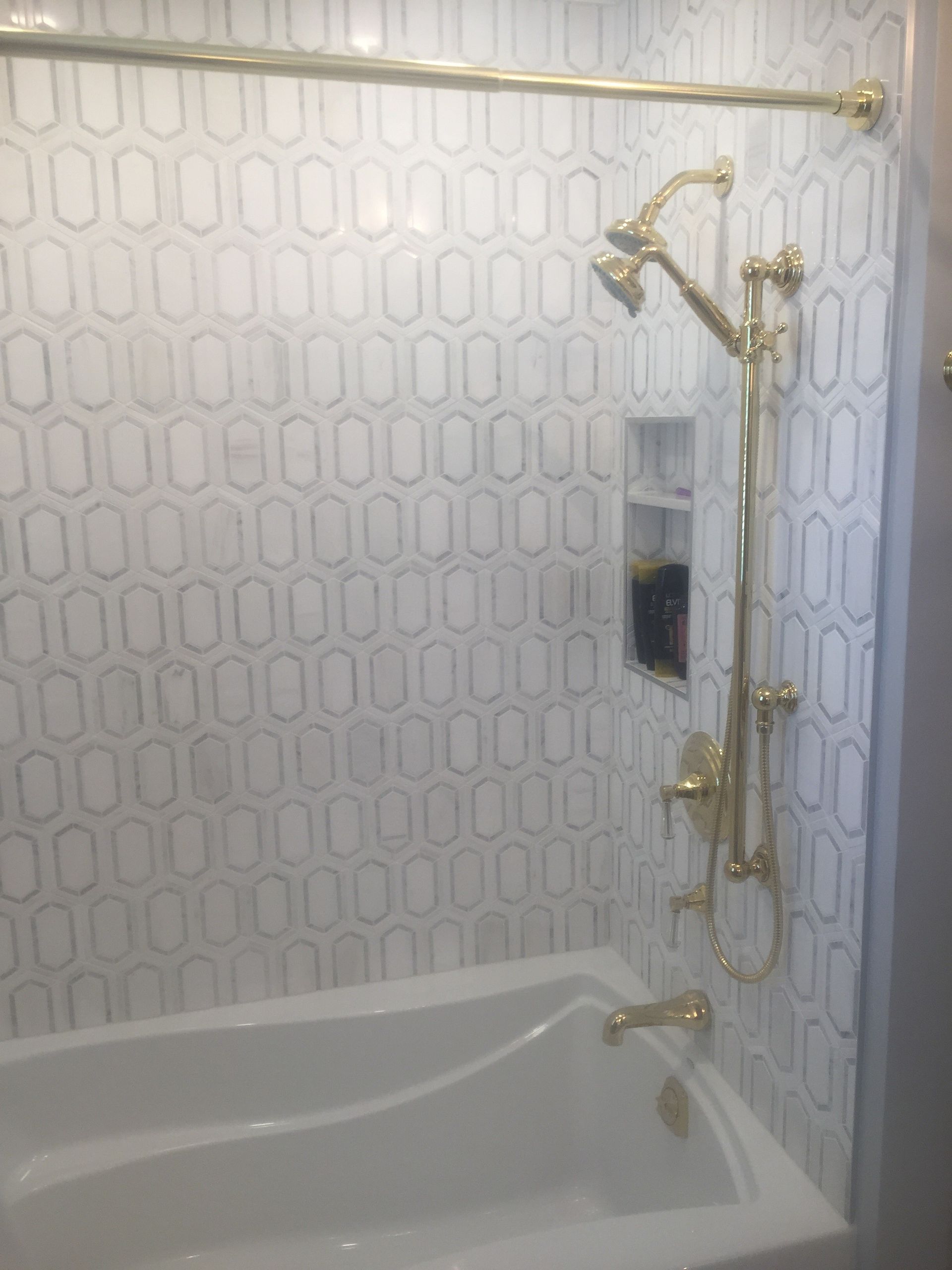 Bathroom with white patterned tile, bathtub, gold grab bars, and a wall-mounted shower hose.