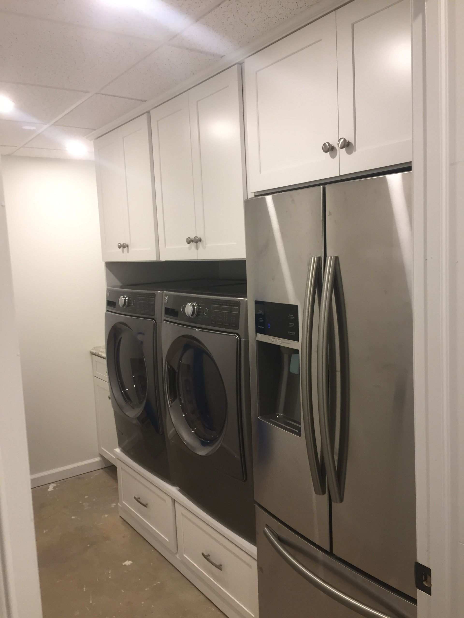 Narrow laundry room with white cabinets, stacked washer and dryer, and stainless steel refrigerator.